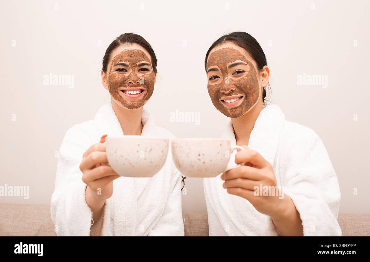 Zwei Freunde mit Schlammmasken auf ihren Gesichtern trinken Tee. Spa-Entspannung, Gesichtshautpflege, Hautreinigung und -Behandlung. Teil einer Serie mit Schlammmaske Stockfoto