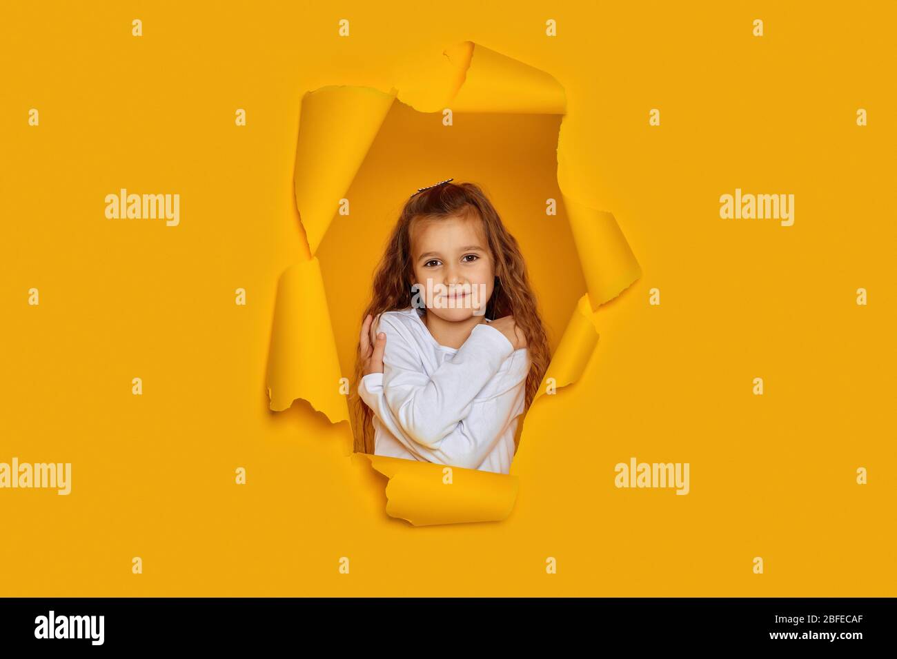 Porträt von niedlichen glücklich lächelnd kaukasischen kleinen Mädchen posiert in gelben Papierloch. Nettes Mädchen träumt von etwas und umarmt sich Stockfoto