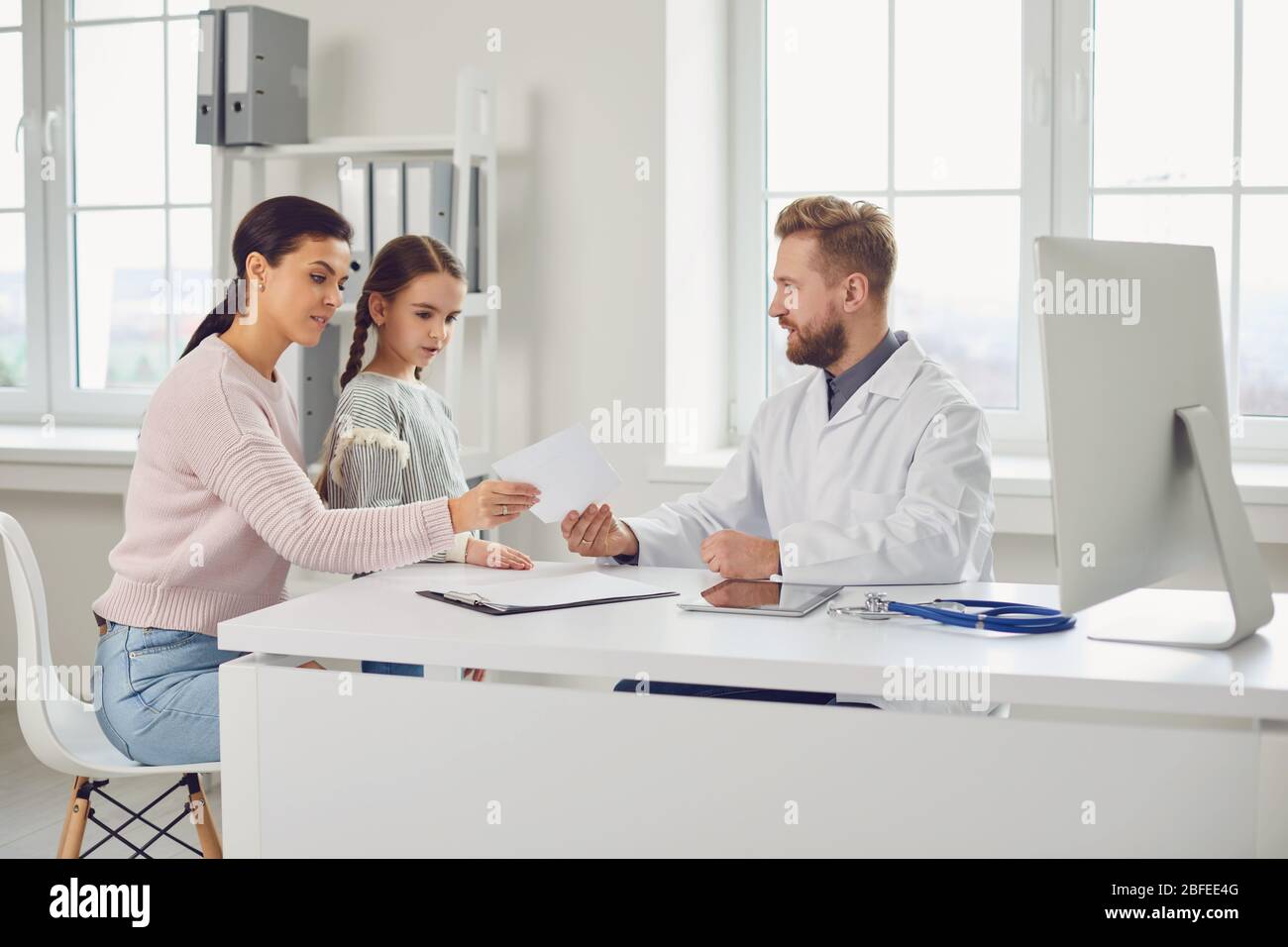 Der männliche Arzt gibt Mutter und Tochter ein Rezept, während er an einem Tisch in einem Klinikbüro sitzt. Stockfoto