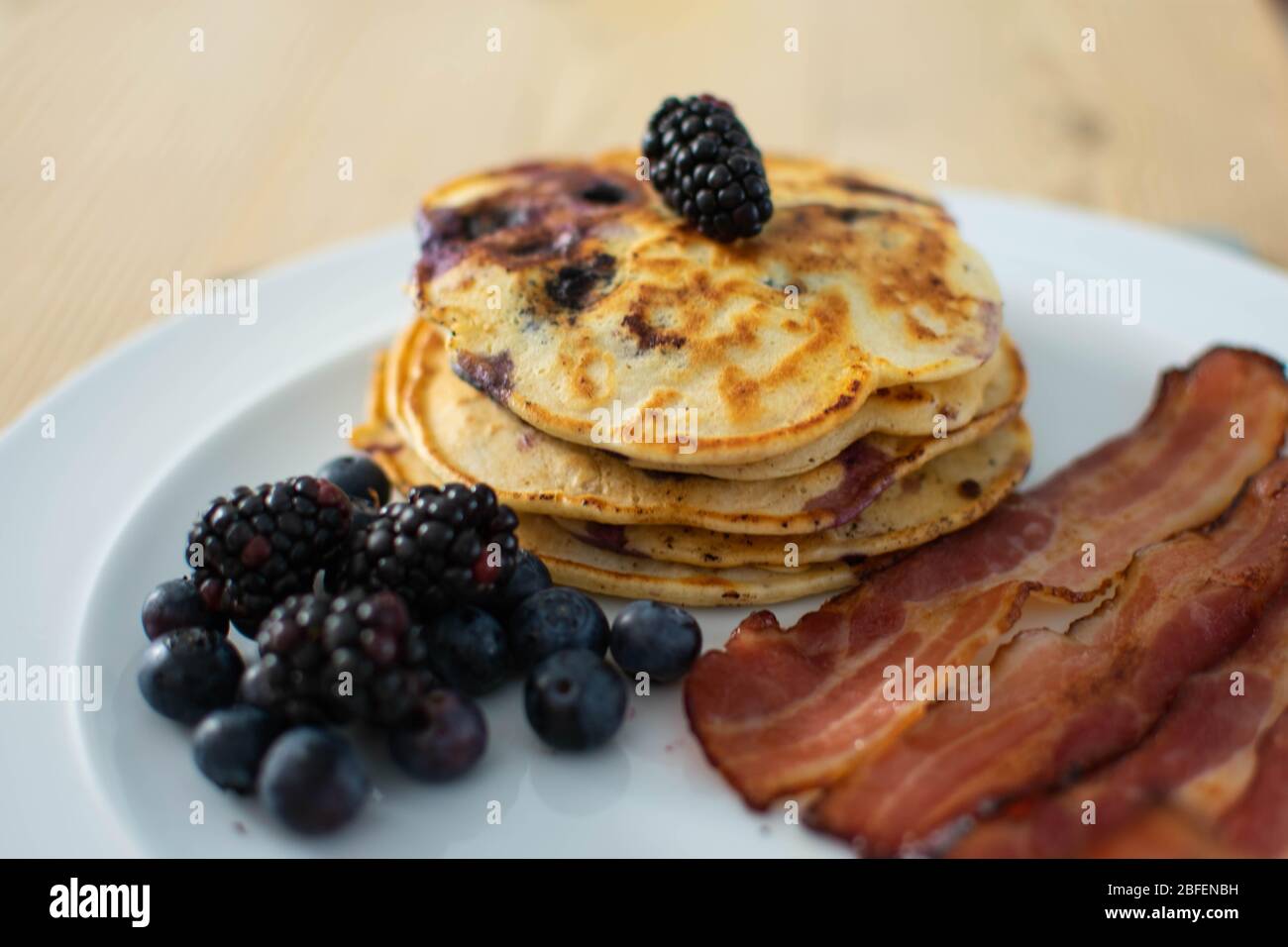 Frühstück mit Heidelbeer-Pfannkuchen mit Speck, Obst und Ahornsirup auf einem weißen Teller serviert. Stockfoto