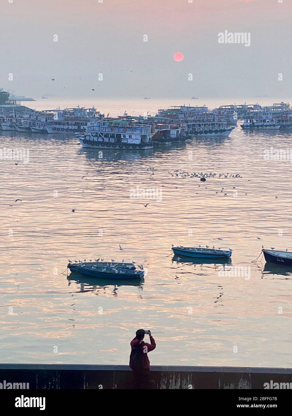 Mann, der den Sonnenaufgang und die Boote durch den Smog in Mumbai fotografiert. Stockfoto