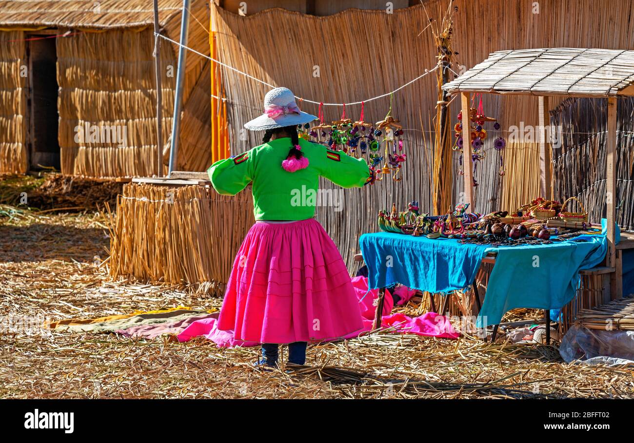 Einheimische Kunst- und Kunsthandwerkskauffrau in auffälligen Stammesfarben auf den schwimmenden Inseln Uros mit Totora-Schilfhäusern und Boden, Titicaca-See, Peru. Stockfoto
