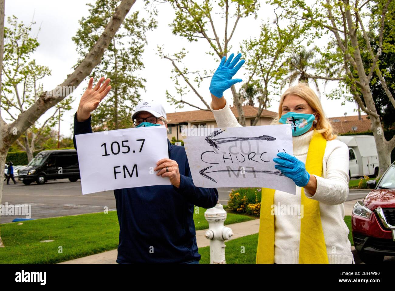 Die Begrüßer bei einem Autofahren Ostern Service in einem Santa Ana, CA, Büropark tragen Sicherheitsmasken wegen des Ausbruchs des Coronavirus oder COVID-19. Notieren Sie sich die Schilder, die den Ort und den Radiosender angeben, an dem die Predigt zu hören ist. Stockfoto
