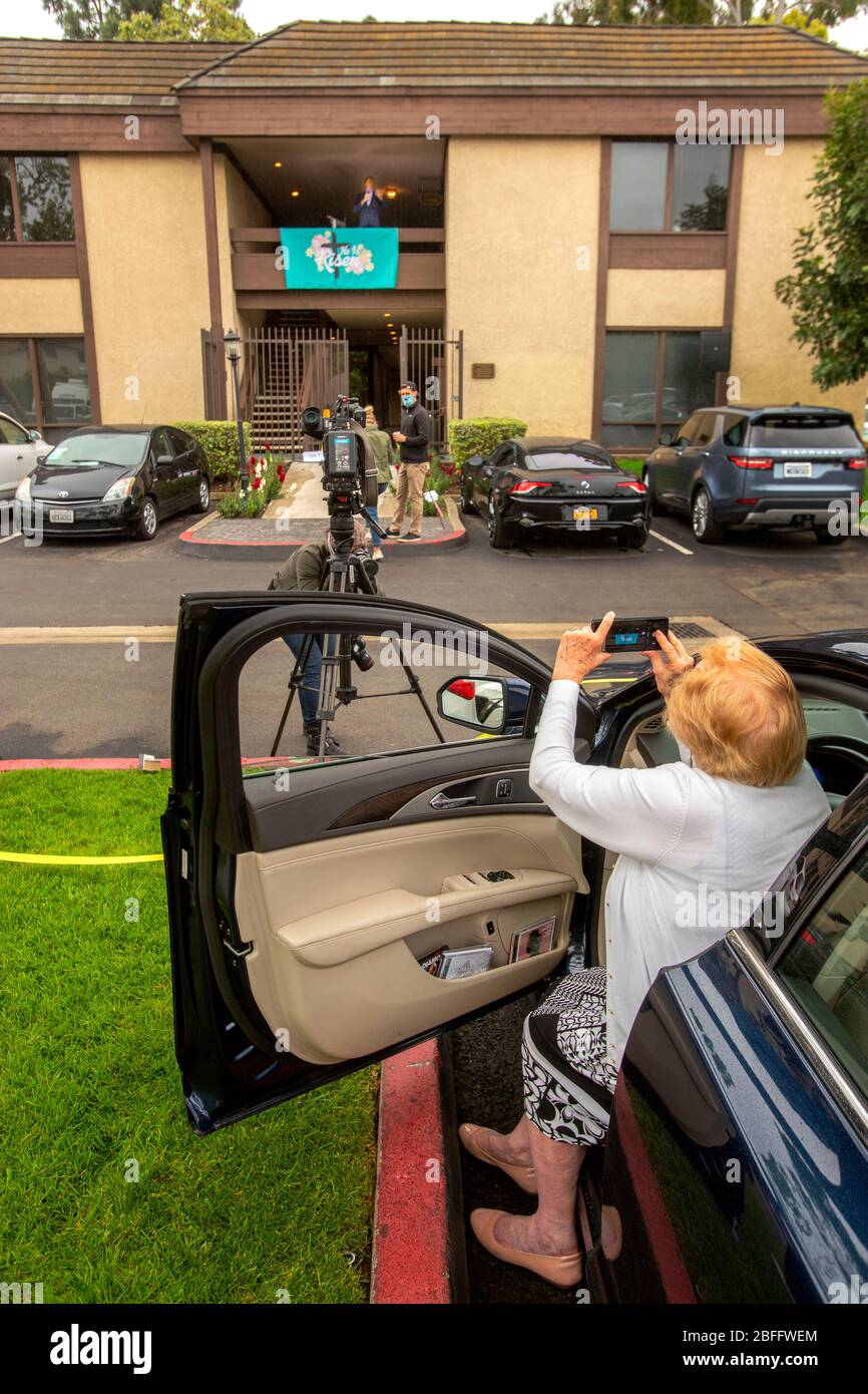 Eine Ministerin bei einem Autofahren-Ostergottesdienst in einem Büropark in Santa Ana, CA, spricht von einem Balkon im zweiten Stock mit einem Spruchband, das sagt: "Er ist auferstanden", während eine Verehrerin ein Bild aus ihrem Auto schnappt. Der Drive-in-Service wurde durch den Ausbruch des Coronavirus oder COVID-19 ausgelöst Stockfoto