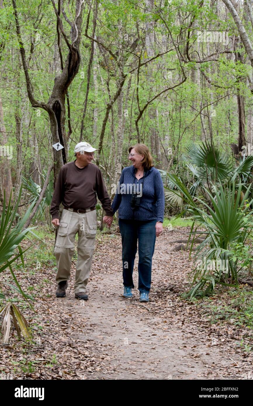 Ein aktives Rentnerpaar, das einen Pfad in einem Wald mit Bäumen und Palmen, die sich gegenseitig betrachten, begibt Stockfoto