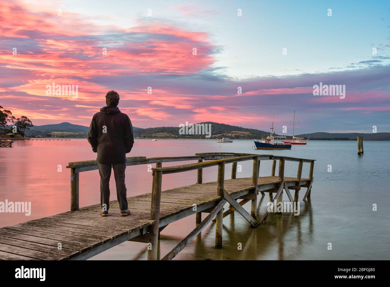 Touristen stehen auf einem alten hölzernen Steg und sehen den perfekten Sonnenuntergang am Hafen, mit Booten sicher in St Helen''s Bay in Tasmanien verankert. Stockfoto