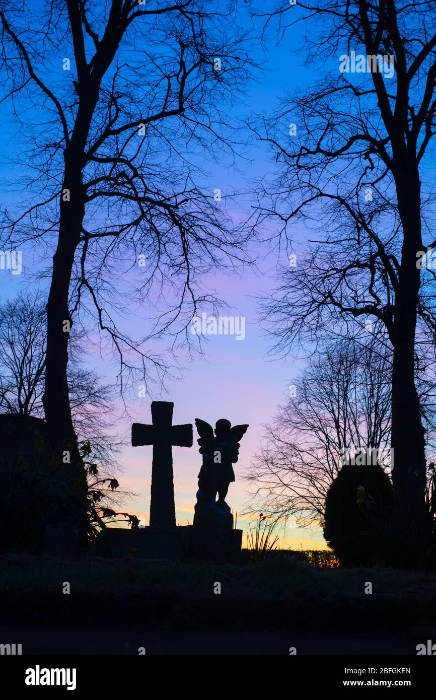 Die Silhouette des Engels und des Kreuzkopfes mit den Bäumen im Friedhof in der Morgendämmerung. Kings Sutton, Northamptonshire, England. Silhouette Stockfoto
