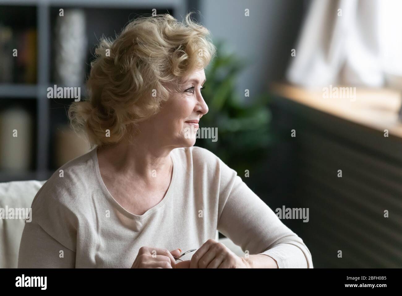 Lächelnde ältere Frau, die auf dem Sofa sitzt und aus dem Fenster schaut Stockfoto