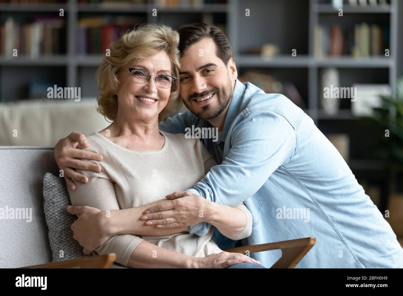 Erwachsener Sohn umarmt ältere Mutter, während sie auf Sessel ruht Stockfoto
