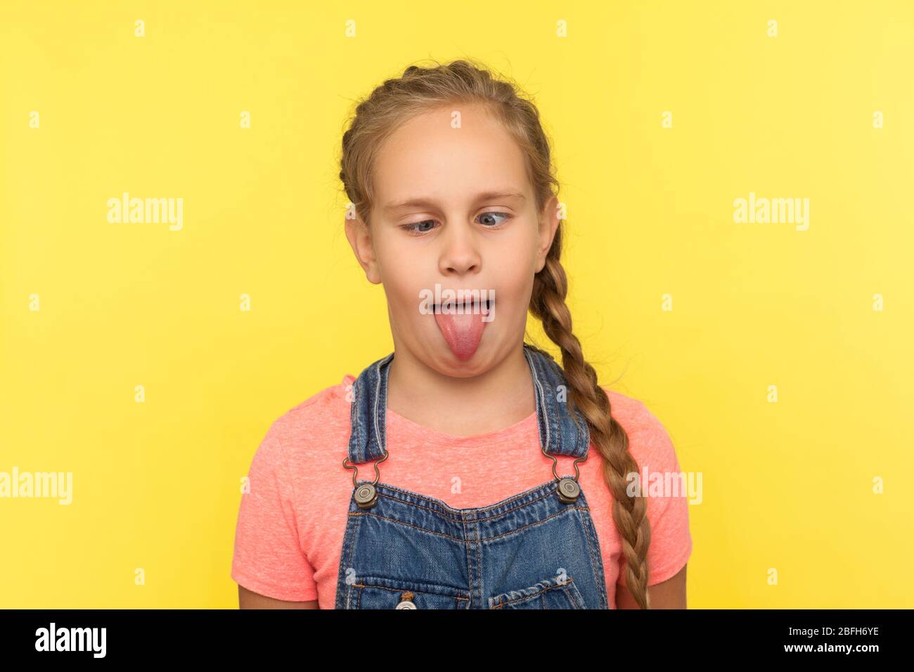 Portrait von entzückenden lustigen kleinen Mädchen in Denim Overalls, die herumtöricht, dumme Gesicht mit gekreuzten Augen und herausstehende Zunge, suchen dumm stu Stockfoto
