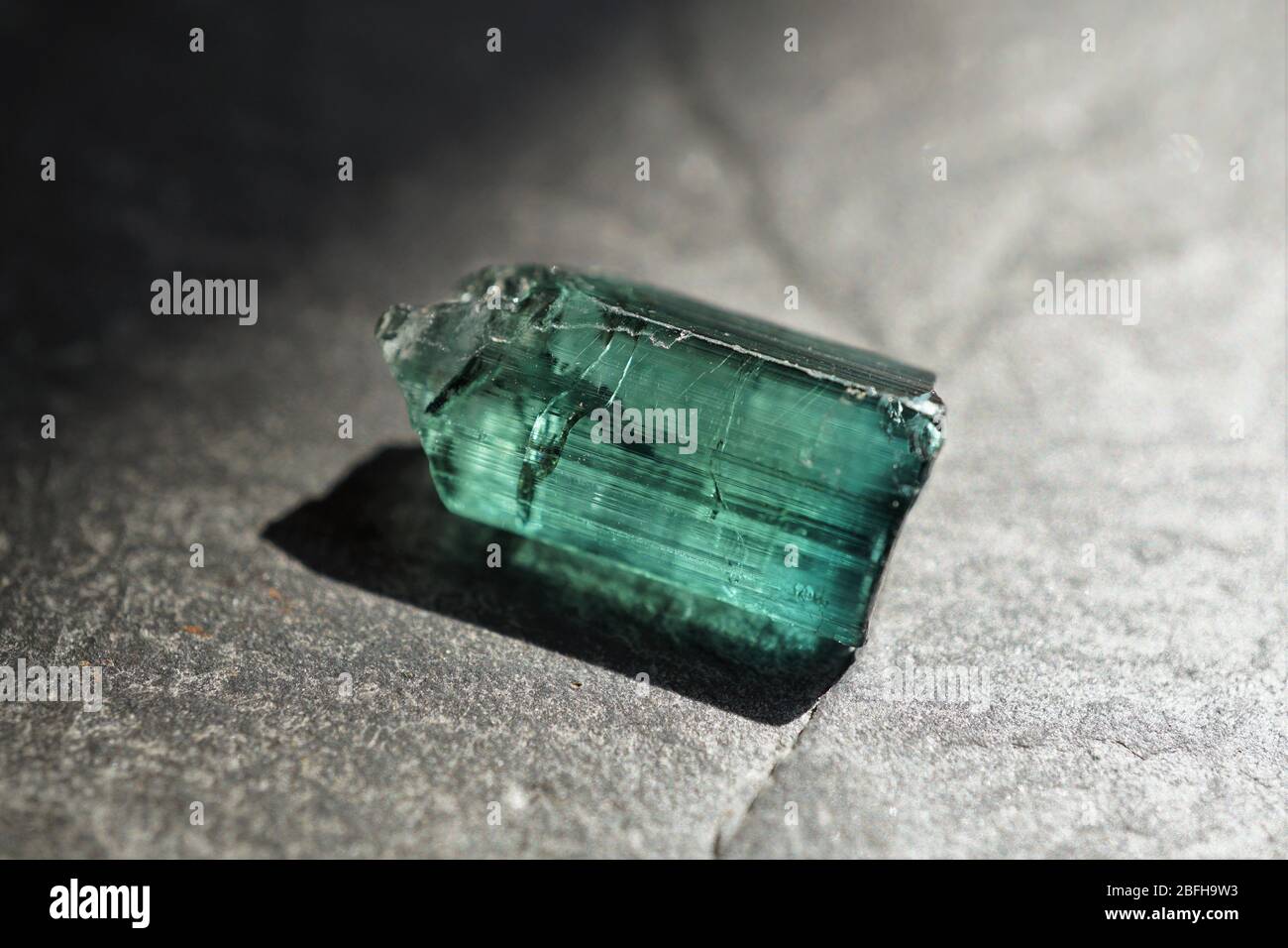 Blau-grüner Turmalin-Rohkristall, auf grauem Schiefergrund, von der Sonne beleuchtet Stockfoto