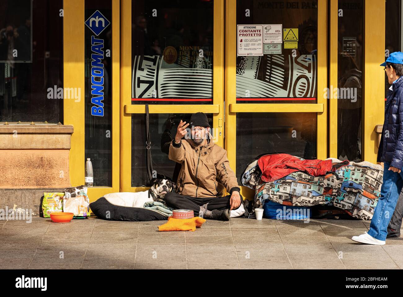 Ein Obdachloser mit seinem Hund bittet an einem Wintersonntag vor einer Bank in der Via Rizzoli, in der Innenstadt von Bologna, um Almosen. Emilia-Romagna, Italien, Europa Stockfoto