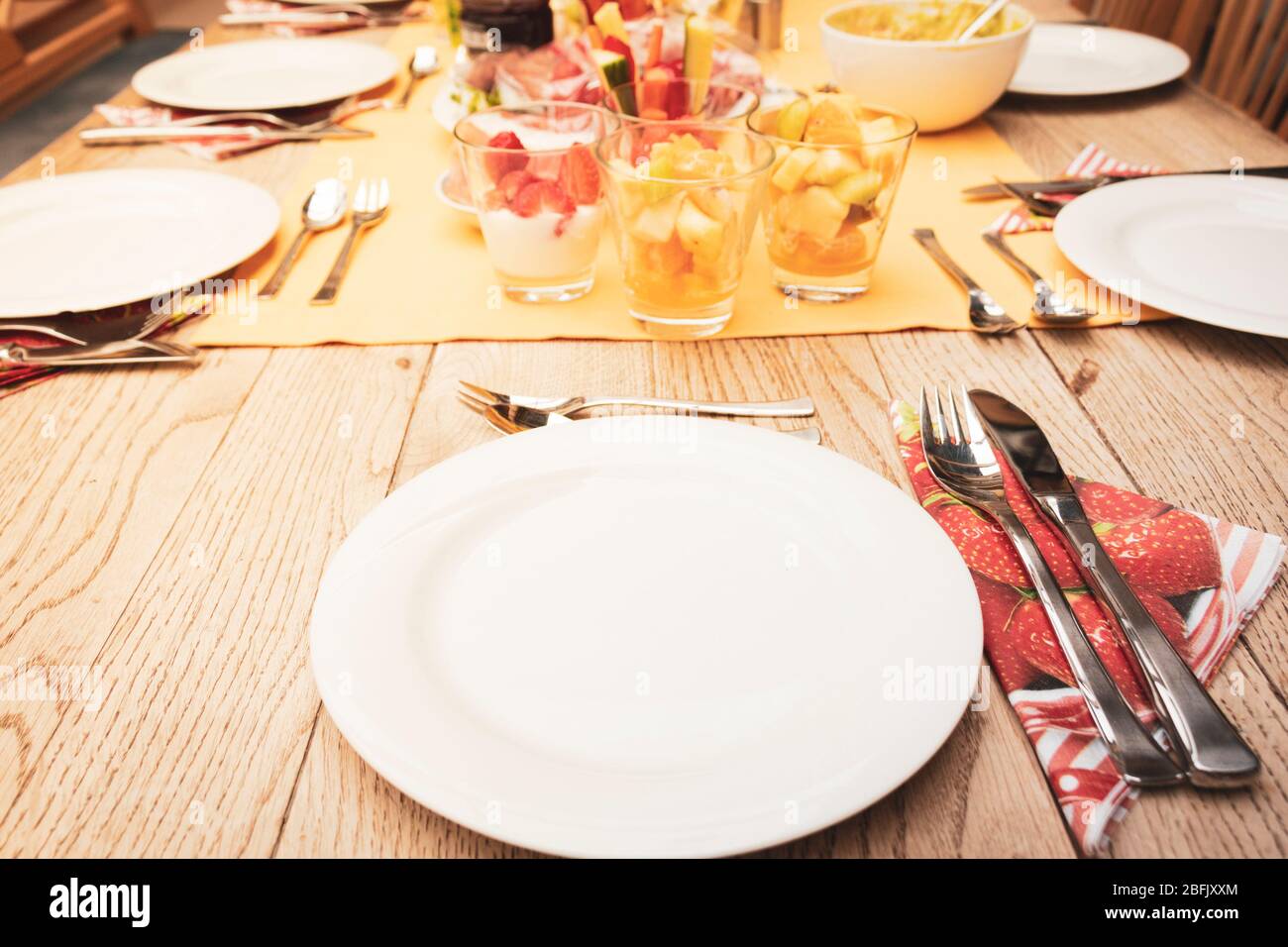 Echter Frühstückstisch mit kleinen Glasschalen voller Früchte und Fisch, gelber Tischdecke, silbernem Besteck, weißen Tellern auf Holztisch Stockfoto