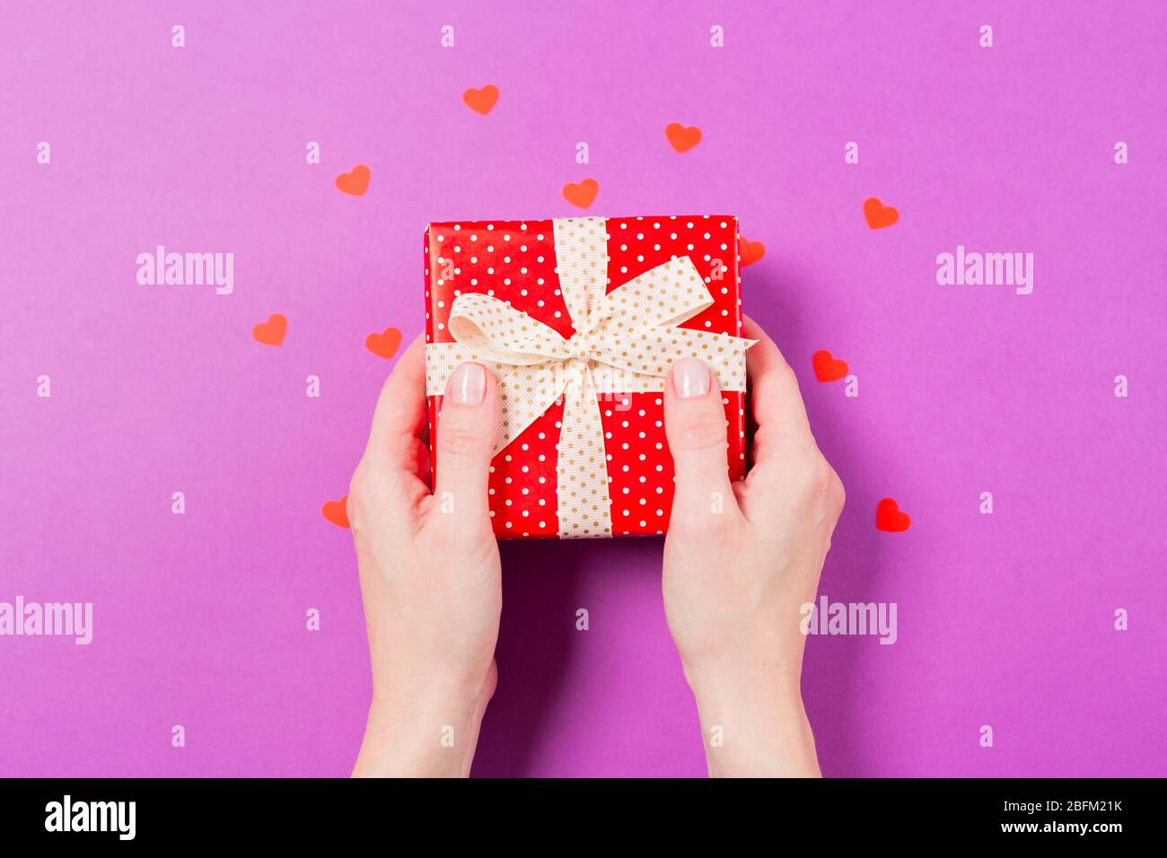 Frau Hände halten rote Geschenk-Box mit kleinen roten Herzen um auf lila Hintergrund. Konzept des Muttertages. Konzept zum Valentinstag. Urlaub Stockfoto