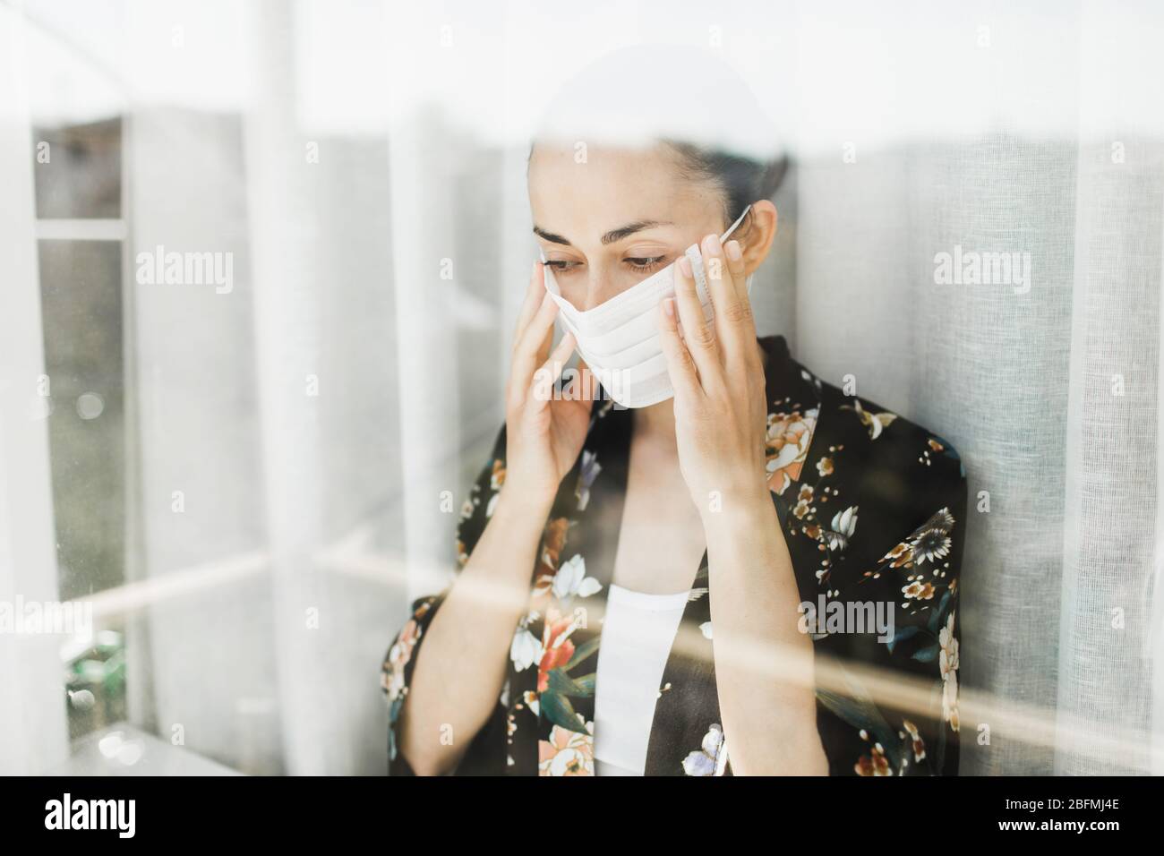 Selbstisolierung in der Quarantäne des Coronavirus. Frau mit medizinischer Maske. Blick durch das Fenster. Verhinderung der Ausbreitung von Viren. Stockfoto