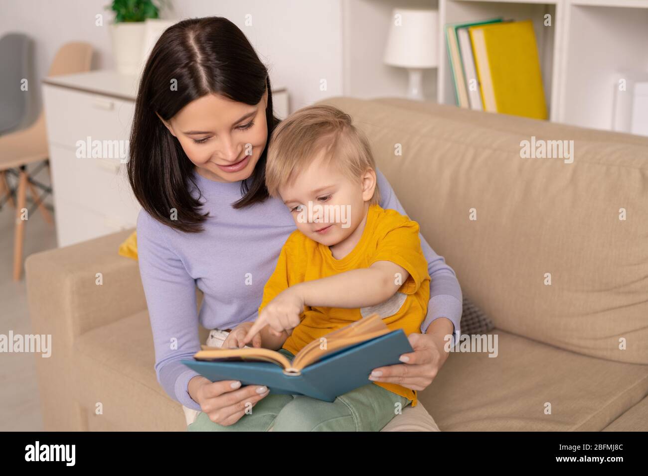 Niedlicher kleiner Junge zeigt auf Bild auf Seite des Buches, während die Diskussion eines der Märchen mit seiner Mutter während der Zeit der Heimat Isolation Stockfoto