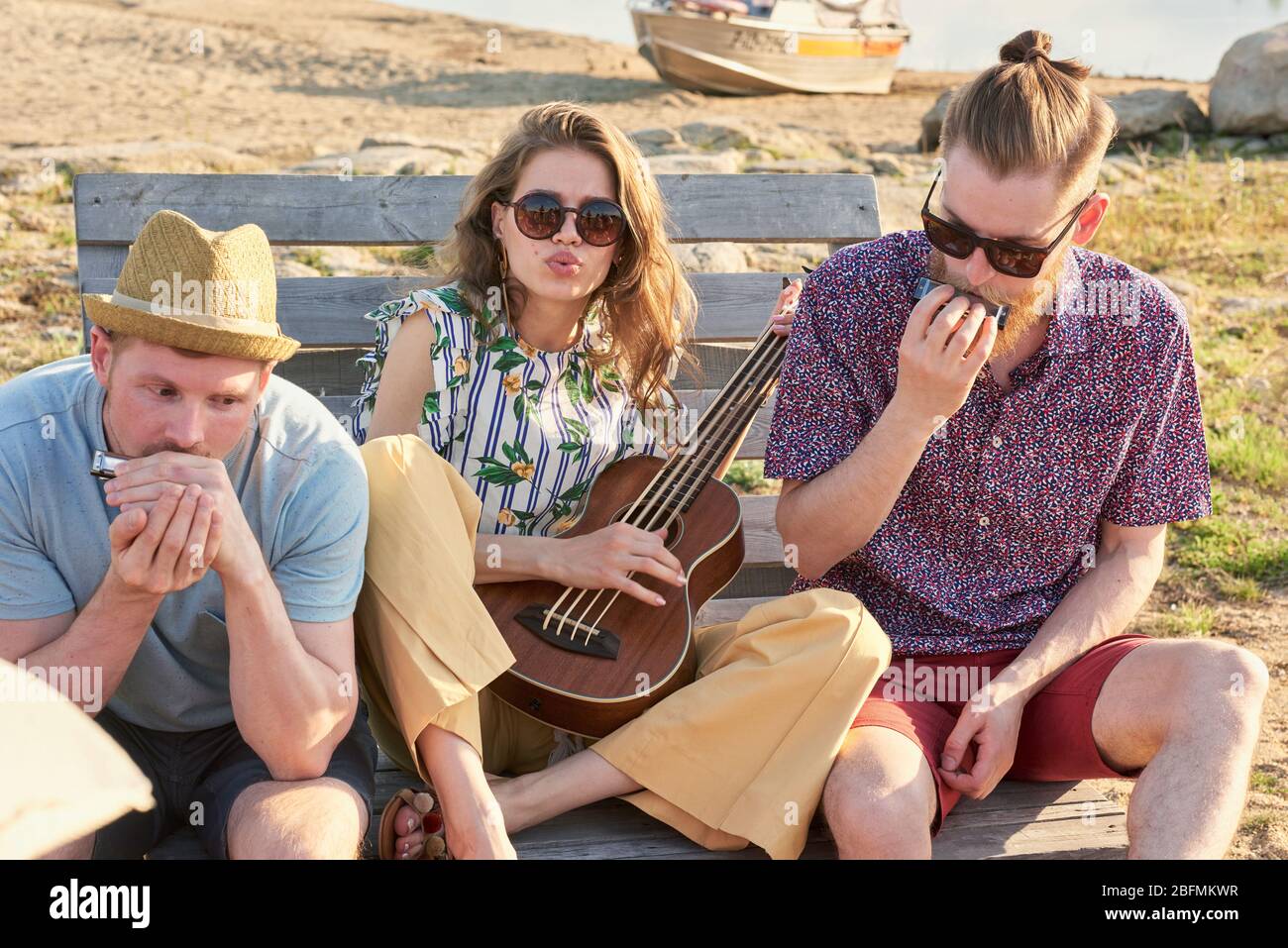 Gruppe von stilvollen jungen Menschen, die sich auf Holzbank am Strand ausruhen und an warmen Sommerabenden Musikinstrumente spielen Stockfoto