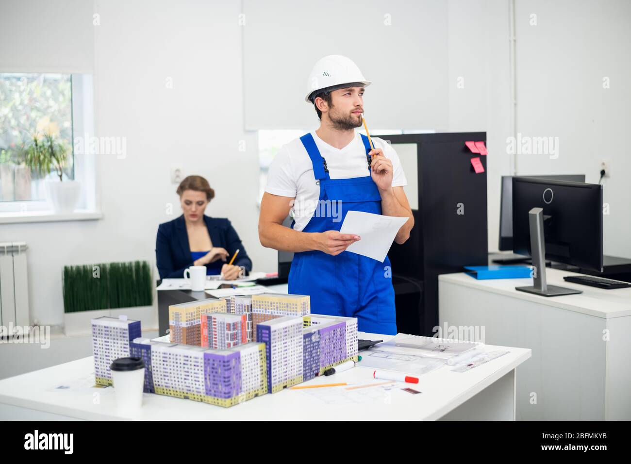 Ein nachdenklicher junger Bauarbeiter im Büro mit einem Baumodell neben ihm. Stockfoto