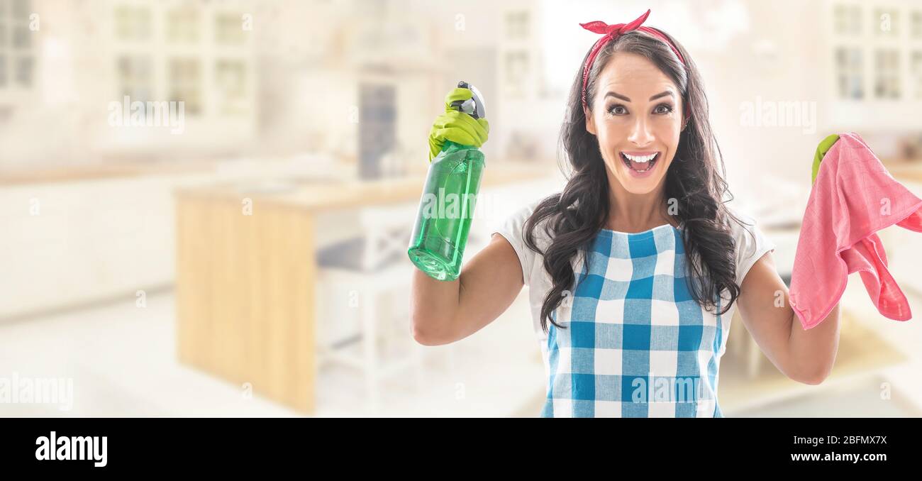 Überraschte junge Frau in der Küche, die in Hauskleidung gekleidet war und ein grünes Waschmittel und ein rosa Reinigungstuch hielt. Stockfoto