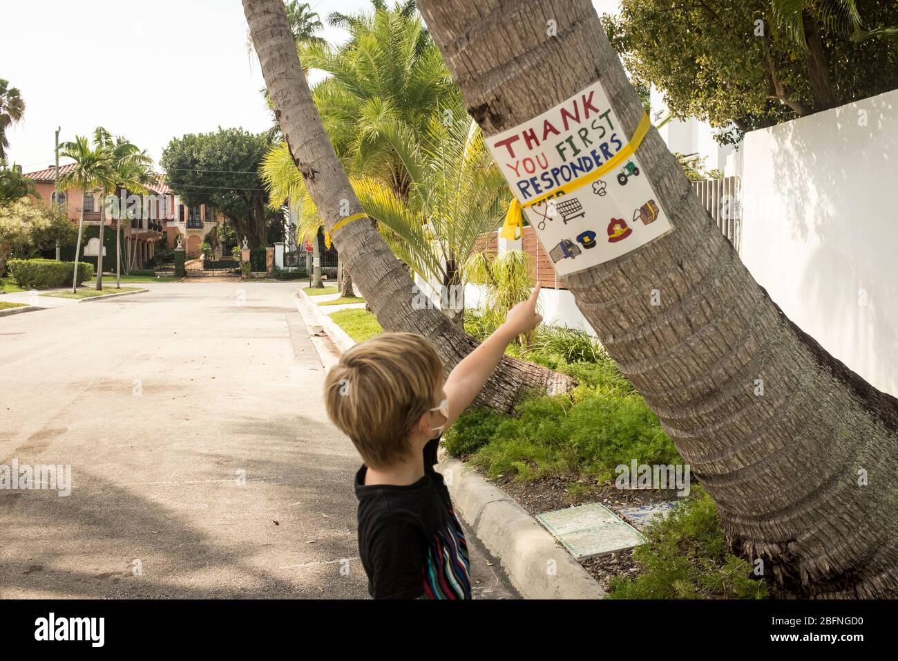 Ein Junge, der während eines Spaziergangs eine Maske trägt, zeigt auf ein selbstgemachtes Schild, das an einer Palme festgeklebt ist, und dankt den Ersthelfern während der Sperrung von COVID-19 in Miami, Florida, USA. Stockfoto