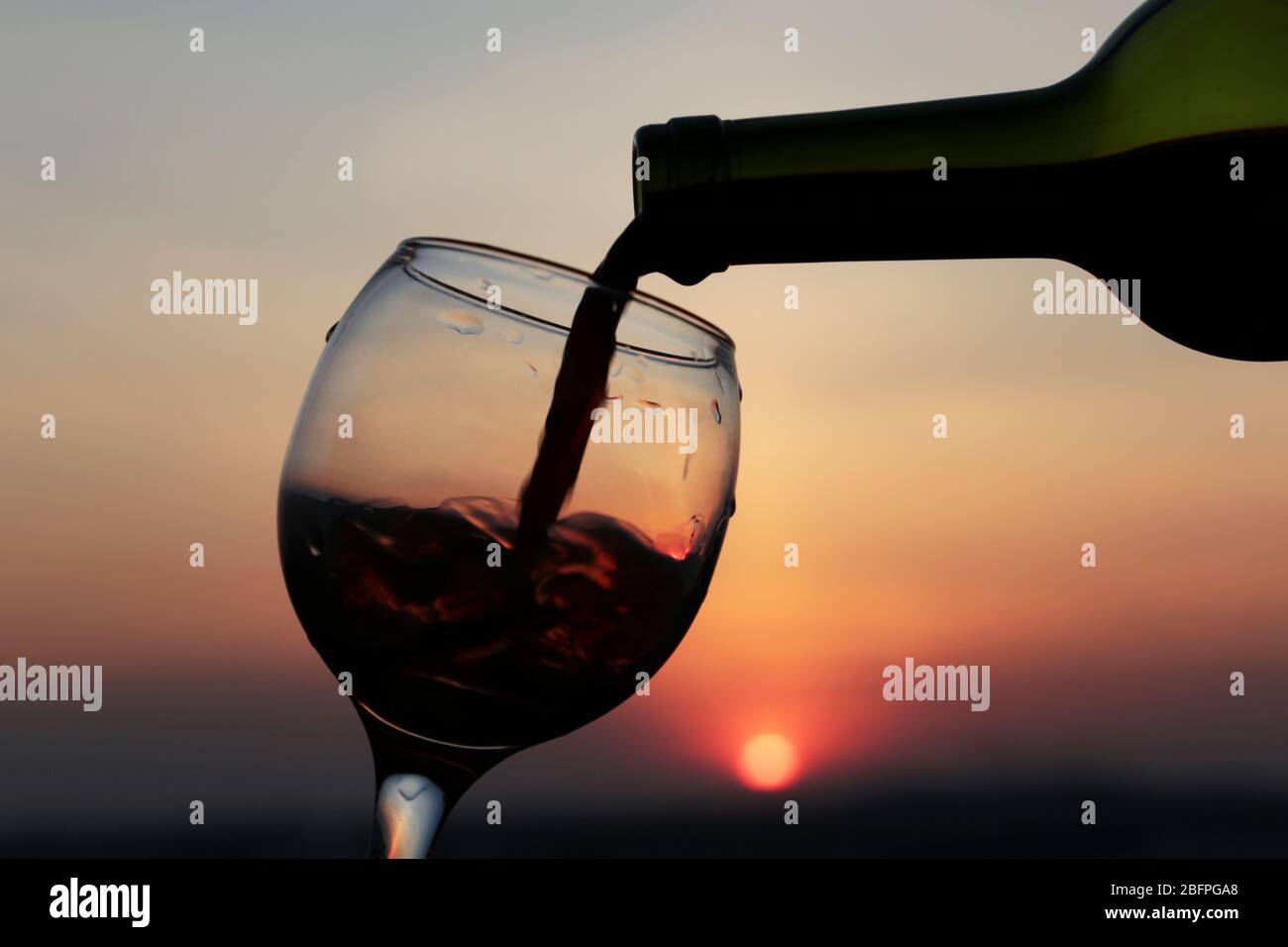 Rotwein aus einer Flasche ins Glas auf wunderschönem Sonnenuntergang Hintergrund scheint die orange Sonne durch die Wolken. Konzept der Feier Stockfoto