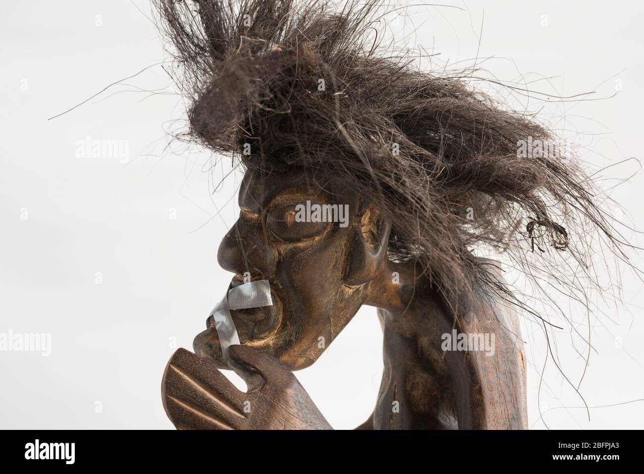 Holz geschnitzte Figur des primitiven Mann trägt Strohrock & lange schwarze zerzauste Haare, aufgeklebt Mund. Konzept; Soziale Unbeweglichkeit, zum Schweigen gebracht, ignoriert. Stockfoto
