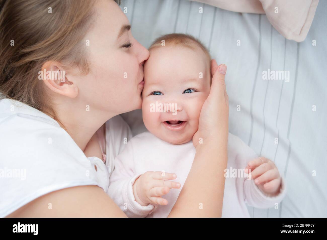 Mutter küsst ihr Baby im Bett. Glückliche Mutter küsst Baby auf dem Bett liegen. Schönes lächelndes Mädchen auf dem Rücken liegend. Draufsicht. Flaches Lay Stockfoto