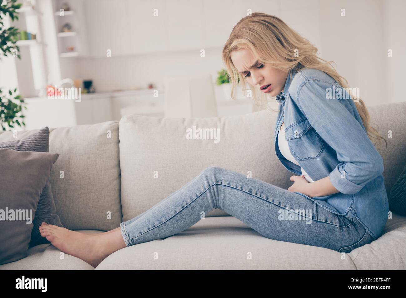 Profilfoto von häuslichen ziemlich unzufrieden blonde Dame sitzende Couch zu Hause bleiben Quarantäne Zeit halten Hände auf Bauch leiden Schmerzen Magen pms Stockfoto