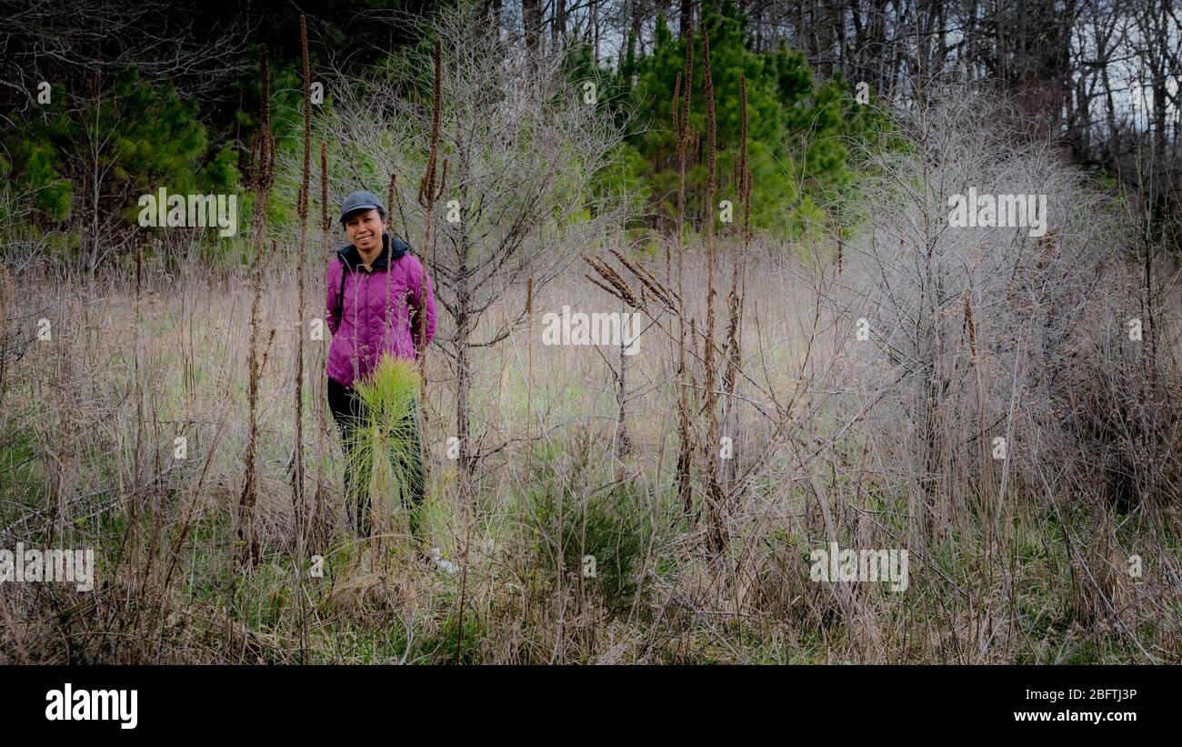 Frau in Mütze und lila Mantel, die inmitten der Bürste in einem Wald neben einem Pfad in Orange County, North Carolina steht Stockfoto