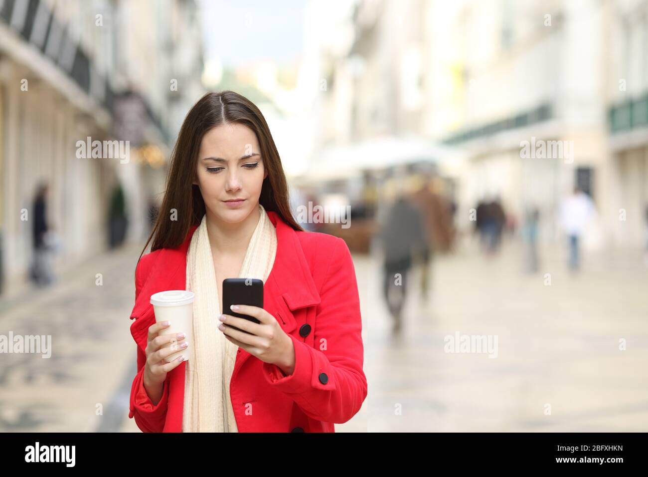 Vorderansicht einer ernsten Frau in rotem Mantel mit Smartphone mit Kaffee zum Mitnehmen, die im Winter auf der Straße spazieren geht Stockfoto