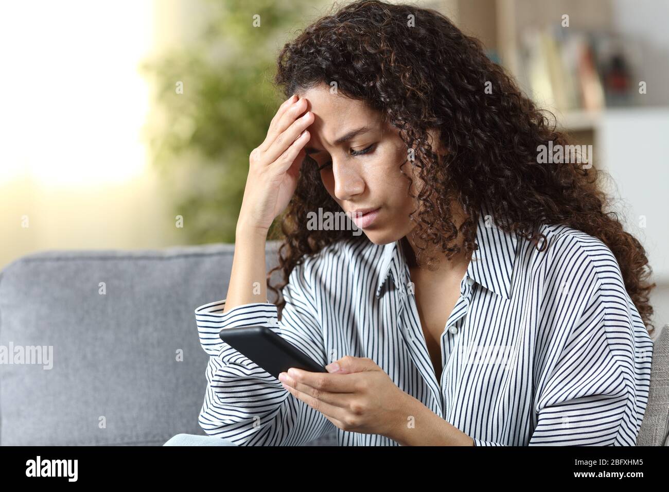 Besorgte lateinische Frau, die auf Smartphone schlechte Nachrichten liest, die zu Hause auf einer Couch sitzen Stockfoto