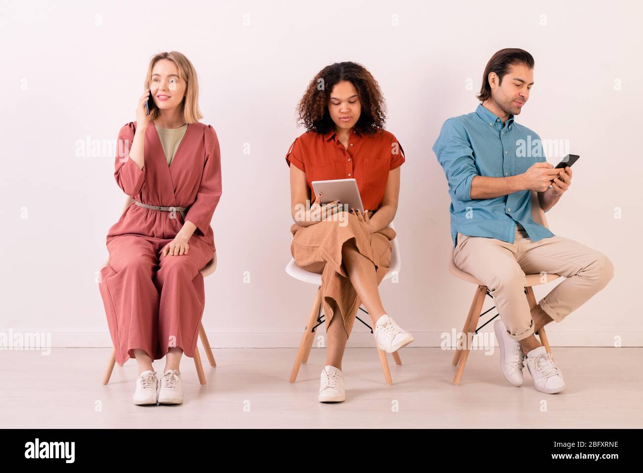 Moderne multi-ethnische Menschen in lässigen Outfits sitzen auf Stühlen in Reihe und mit Gadgets alle Zeit für die Kommunikation Stockfoto