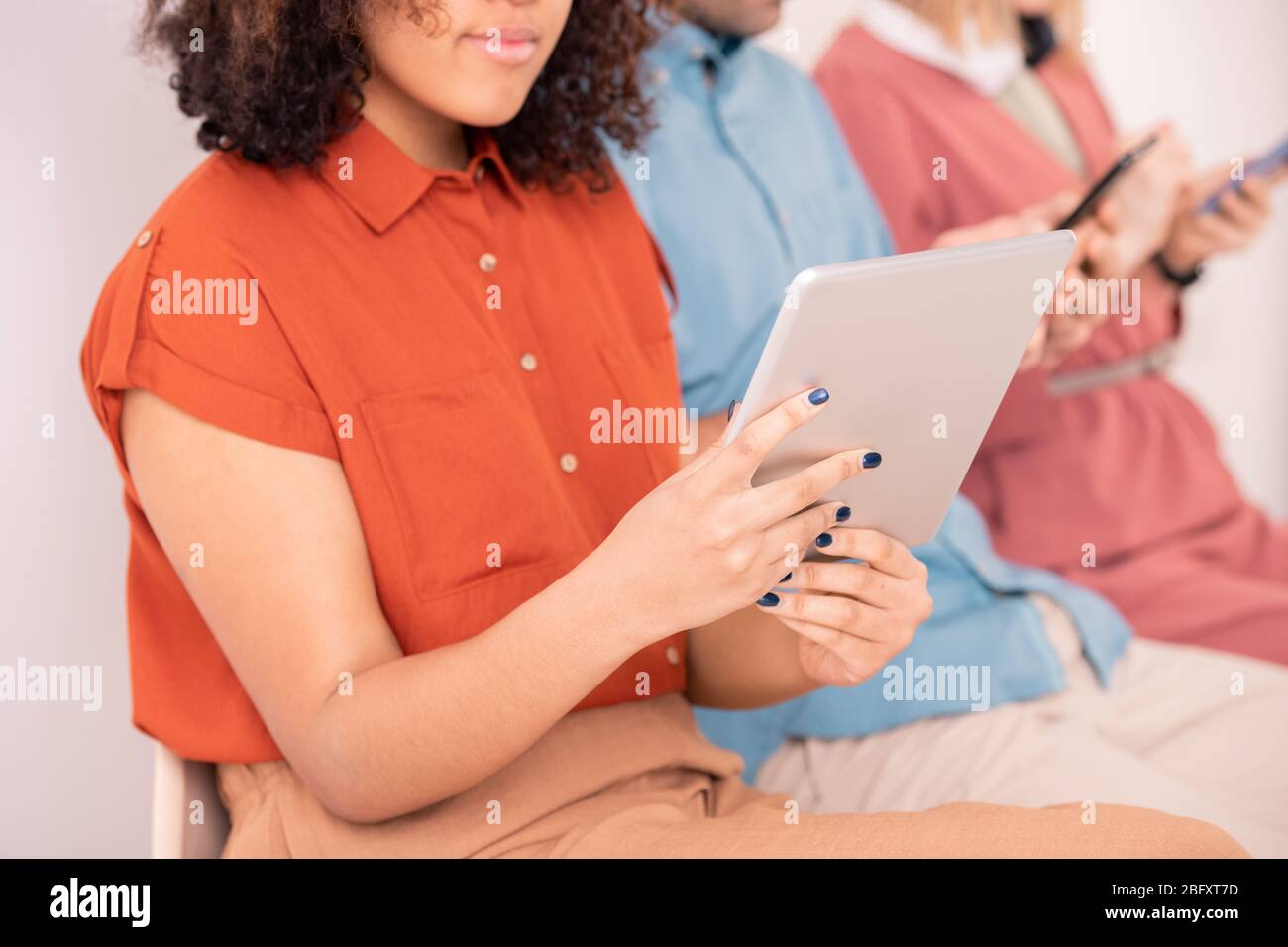 Nahaufnahme einer afroamerikanischen Frau in orangefarbener Bluse, die in einer Reihe von Gadget-abhängigen Menschen sitzt und mit einem Tablet Zeit verschwendet Stockfoto