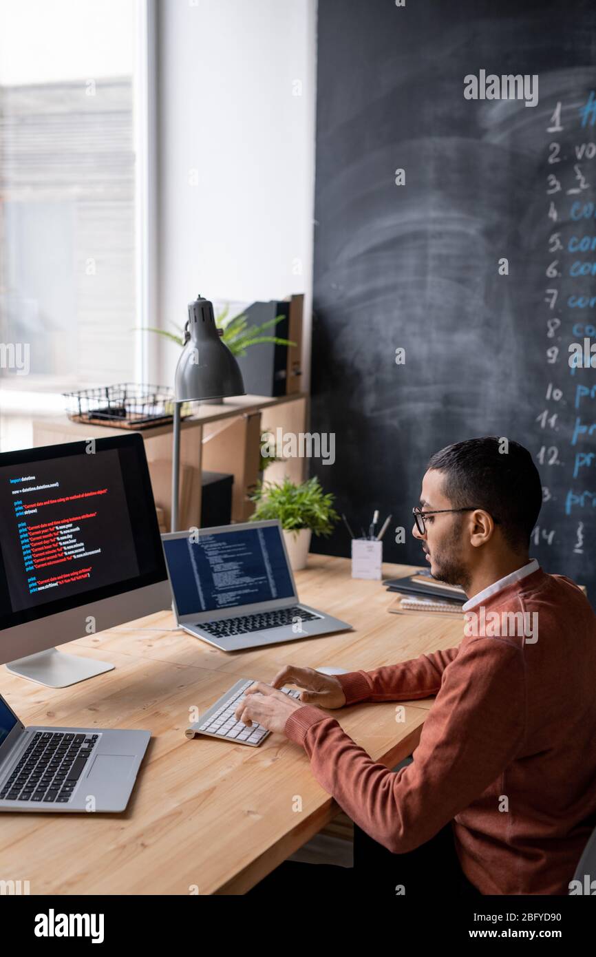 Konzentrierter junger arabischer Programmierer in Brillen, die am Tisch sitzen und auf der Tastatur zwischen drei Computern tippen, während das Skript codiert wird Stockfoto
