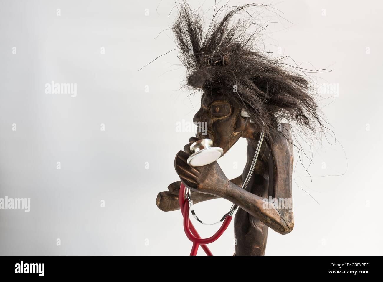 Holz geschnitzte Figur des primitiven Menschen trägt Strohrock & lange schwarze zerzauste Haare und Stethoskop. Konzept; Ehrgeiz, Anspruch, soziale Mobilität. Stockfoto