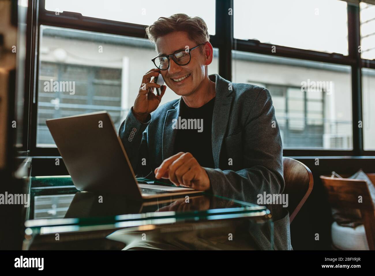 Lächelnder Geschäftsmann mit Laptop, der im Büro auf dem Handy spricht. Ein professioneller Mitarbeiter sitzt am Schreibtisch und telefoniert. Stockfoto