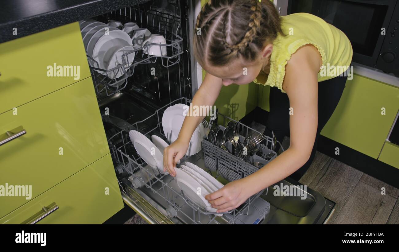 Schlaue Mädchen lernen, Geschirrspüler zu benutzen. Stilvolle, moderne Einbauküche in grünem Schwarz. Kind legt sauberes Geschirr. Stockfoto