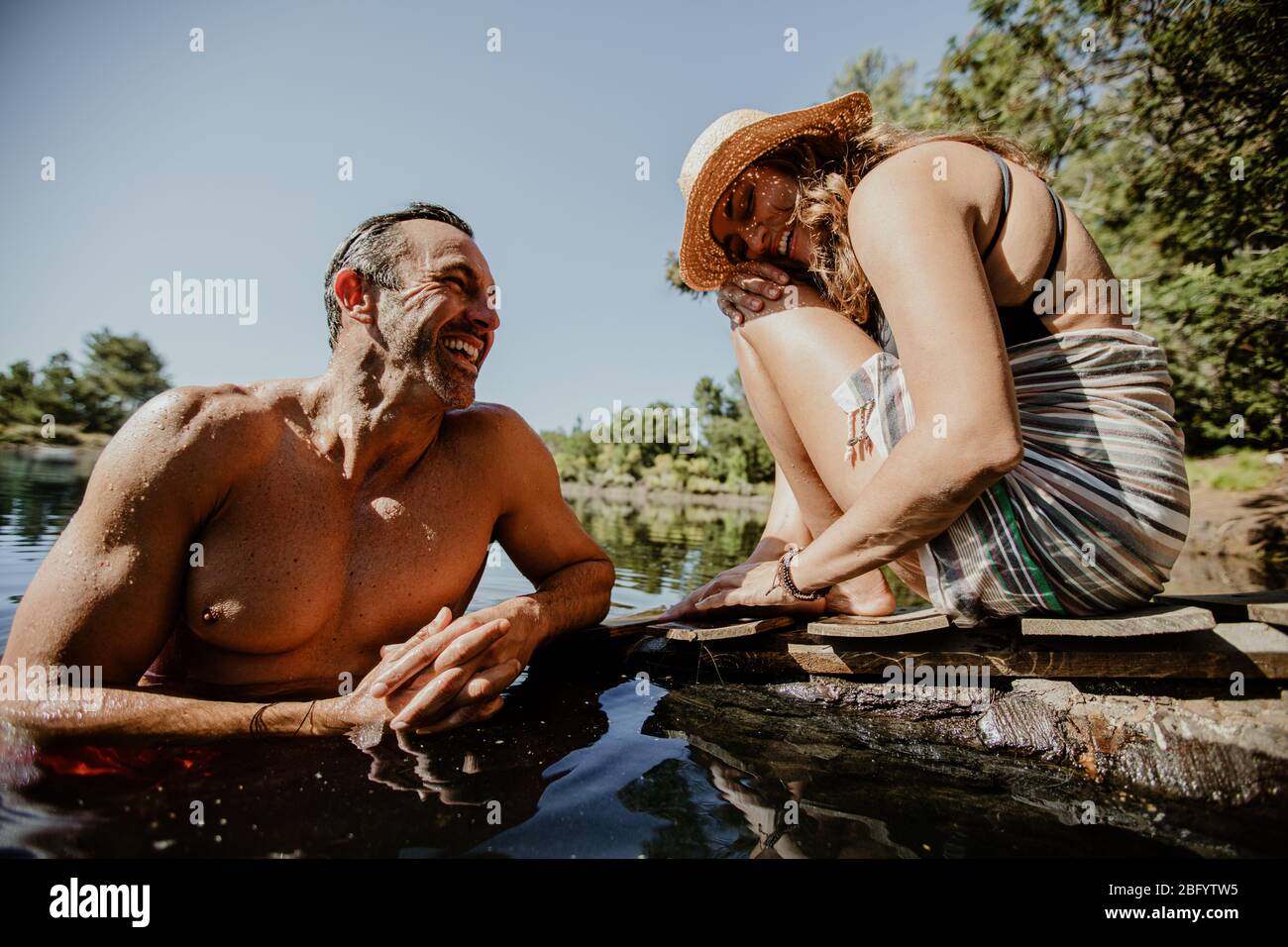 Reifer Mann im See mit Frau am Pier sitzend. Pärchen genießen ihren Camping-Urlaub am See. Stockfoto