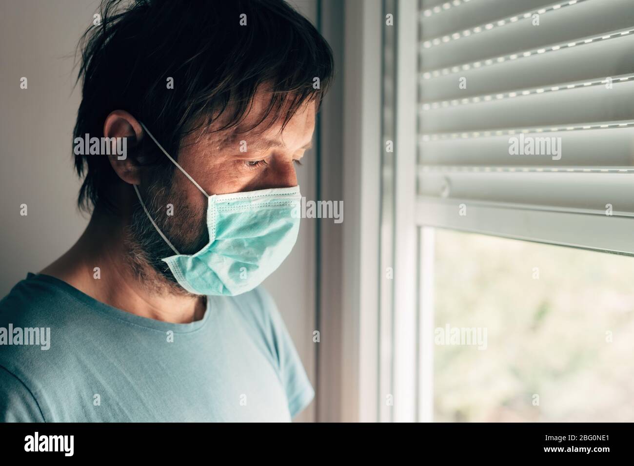 Depressiver Mann mit Schutzmaske in Selbstisolierung Quarantäne während des Virus-Ausbruchs Pandemie, Erwachsener Mann am Fenster stehend Stockfoto