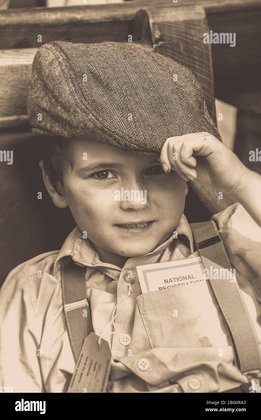 Altmodische Sepia Nahaufnahme von Evakuierten Jungen, vintage Kind in flacher Kappe & Zahnspange, isoliert an vintage Bahnhof, 1940er ww2 Sommerereignis, Großbritannien. Stockfoto