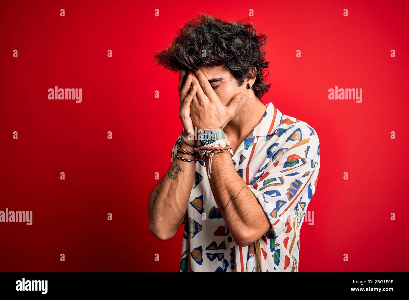 Junger schöner Mann im Urlaub trägt Sommerhirt über isoliertem roten Hintergrund mit trauriger Expression, die Gesicht mit Händen während des Schreiens bedeckt. Depression Stockfoto