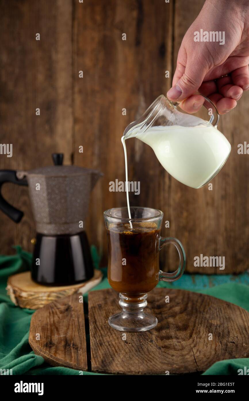 Milch wird in den Karaffe gegossen. Geyser Kaffeemaschine, Kaffeesahne auf einem Holztisch. Glasbecher mit Cappuccino zur Kaffeebereitung. Kaffee zubereiten, Hand Stockfoto