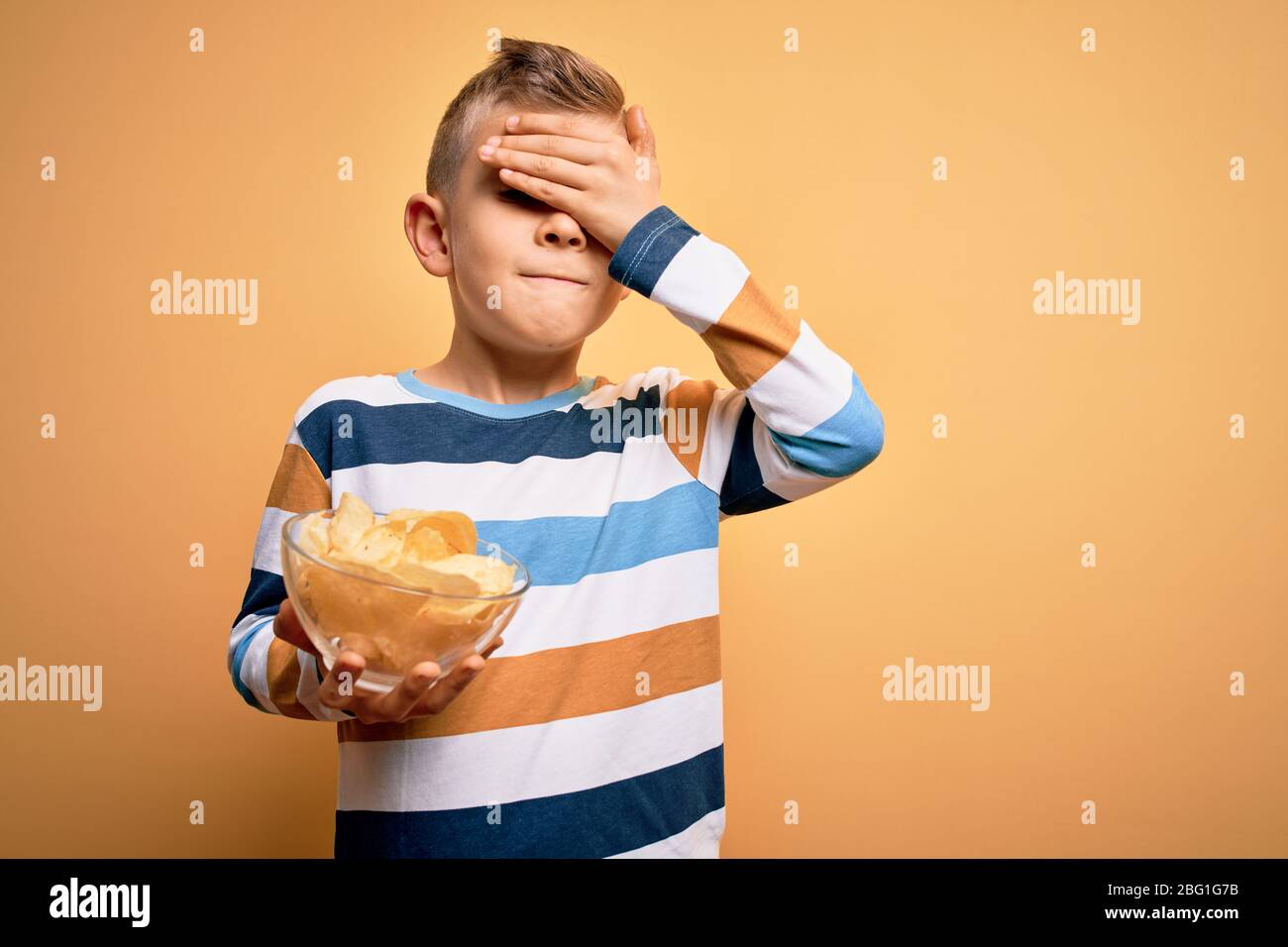 Junge kleine kaukasische Kind essen unheisse Kartoffeln Chips Chips über gelbem Hintergrund gestresst mit Hand auf Kopf, schockiert vor Scham und Überraschung f Stockfoto