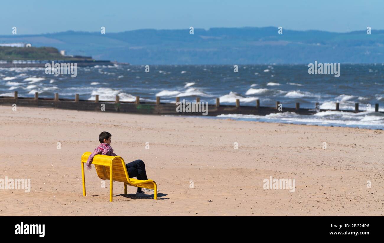 Portobello Strand und Promenade in der Nähe von Edinburgh während der Sperrung des Coronavirus am 19. April 2020. Frau sitzt allein auf der gelben Bank am leeren Strand. Stockfoto