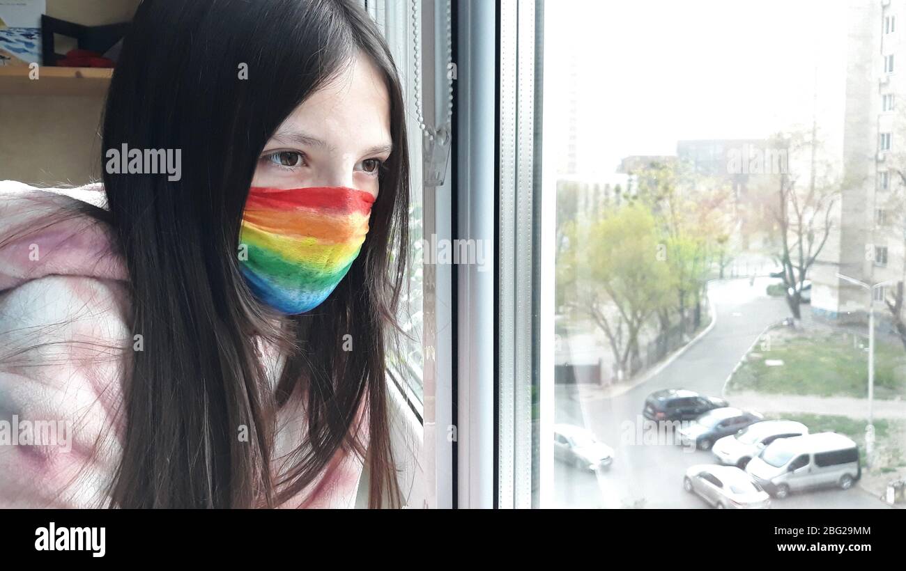 Brunette Teenager-Mädchen in einer medizinischen Maske in hellen Regenbogenfarben gemalt steht am Fenster mit ihrer Hand auf glass.Concept zu Hause bleiben, bleiben Stockfoto