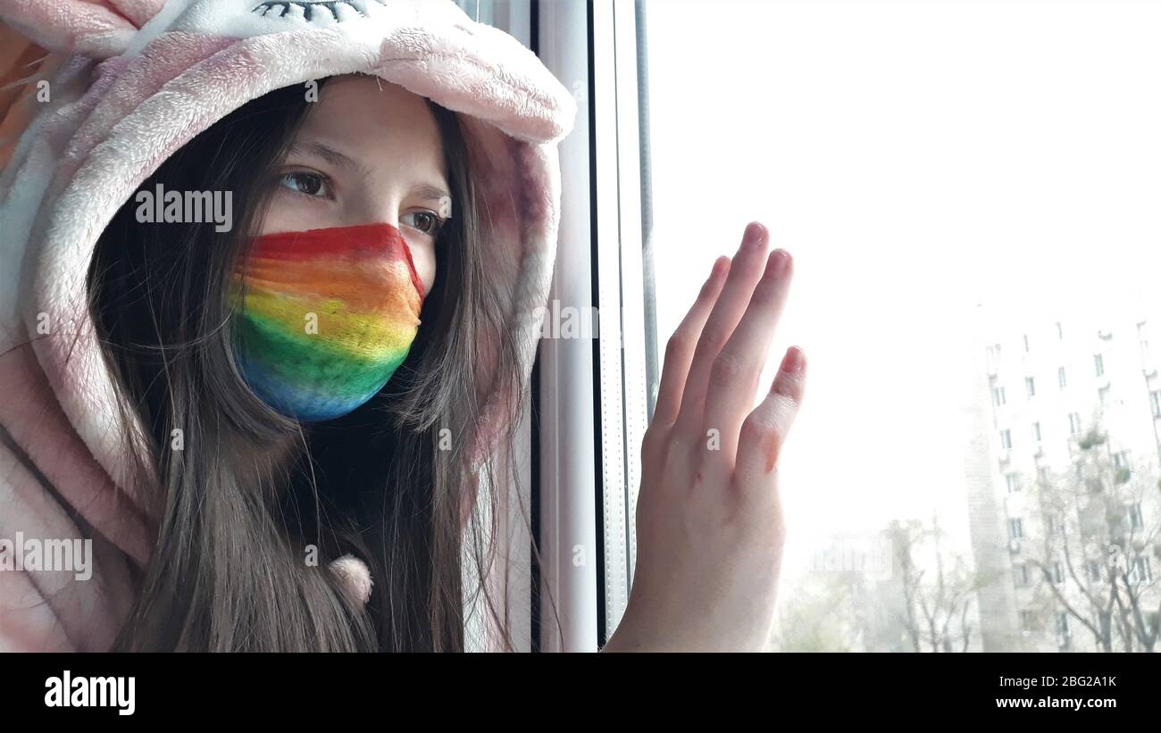 Brunette Teenager-Mädchen in einer medizinischen Maske in hellen Regenbogenfarben gemalt steht am Fenster mit ihrer Hand auf glass.Concept zu Hause bleiben, bleiben Stockfoto