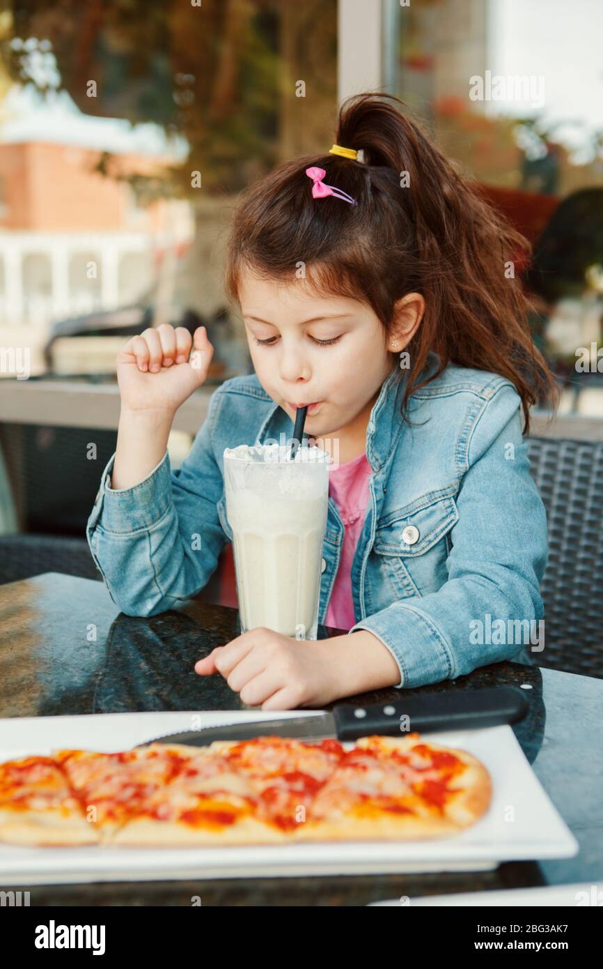 Kaukasische Vorschule Mädchen trinken Milch schütteln mit Papier Stroh im Café. Kind Kind Spaß im Restaurant Terrasse genießen Pizza Snack Essen und Trinken. H Stockfoto
