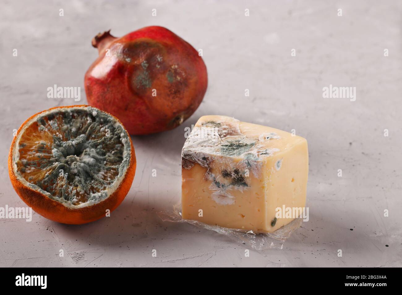 Verdorbene faule Speisen mit Schimmel: Granatapfel, eine halbe Orange und Hartkäse auf grauem Hintergrund Stockfoto