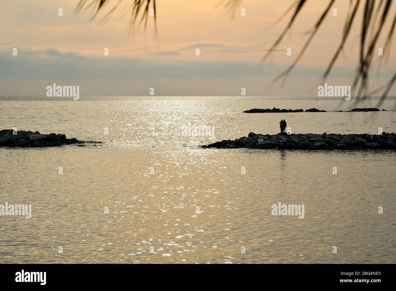 Gegenlicht Seenlandschaft mit der Silhouette eines Paares, das auf einem felsigen Wellenbrecher bei Sonnenuntergang, Sanremo, Imperia, Ligurien, Italien steht Stockfoto