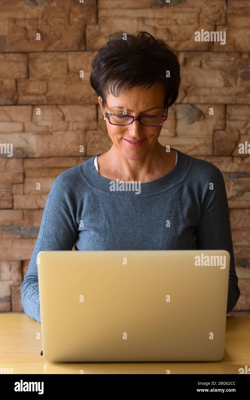 Reife glückliche Frau lächelnd, während das Tragen von Brillen und mit Laptop auf Holztisch gegen Ziegelwand Stockfoto